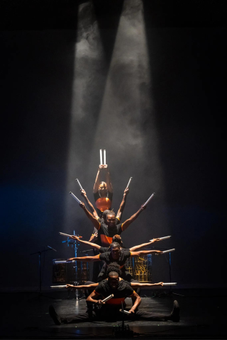 Step Africa! dancers with arms extended, posing as a tower on a dark stage under a single spotlight