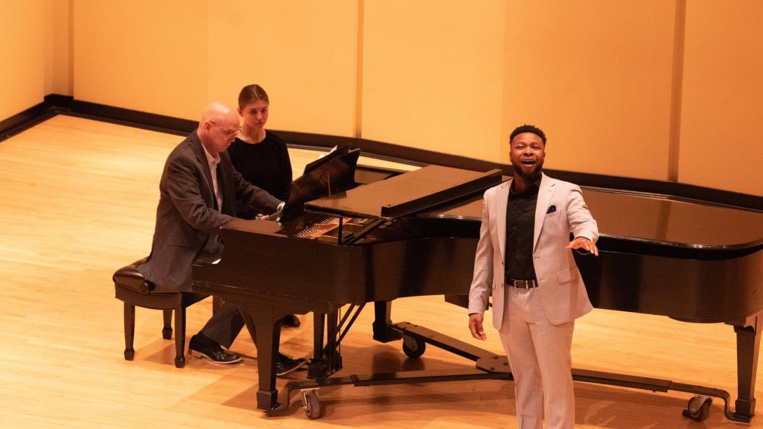 two people sit at a piano; a standing man sings at center
