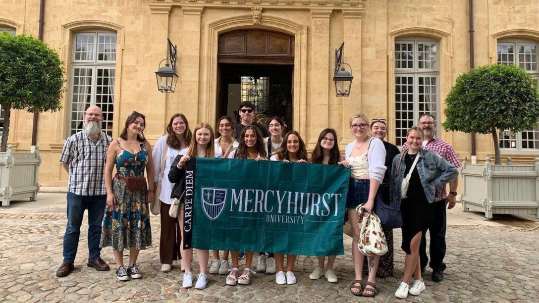 a group of students and professors pose in France with a Mercyhurst flag