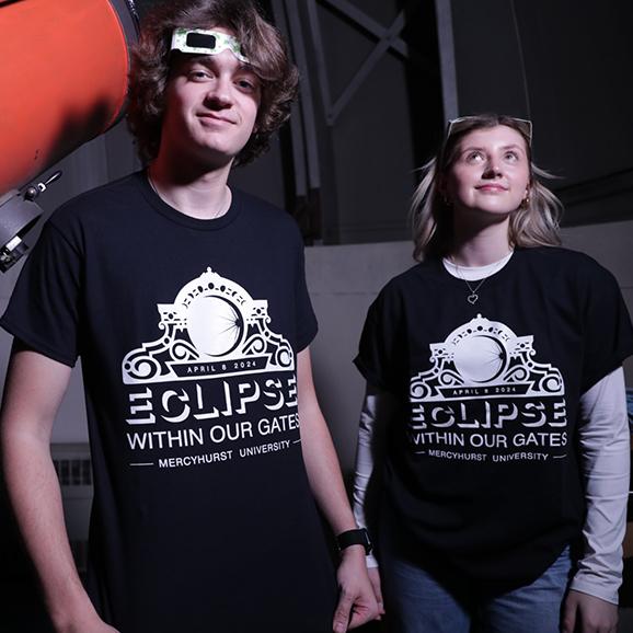 Image of male and female student wearing the 2024 Mercyhurst University Eclipse Within Our Gates t-shirt