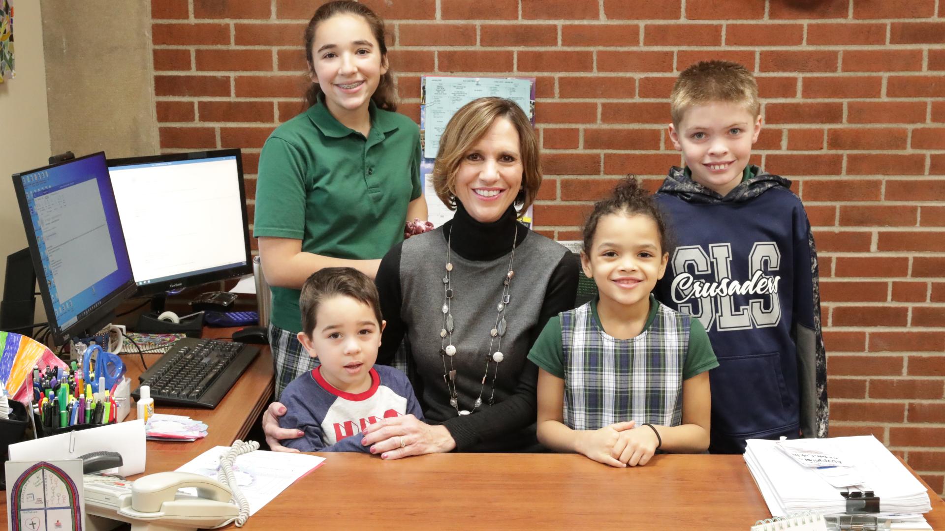 a principal sits at a desk surrounded by four children