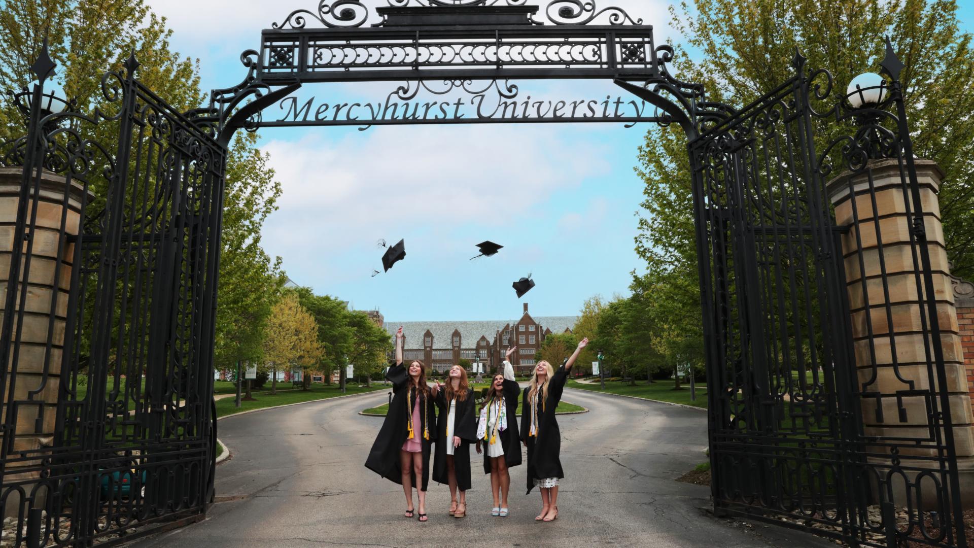 four female students throw graduation caps under the mercyhurst gates