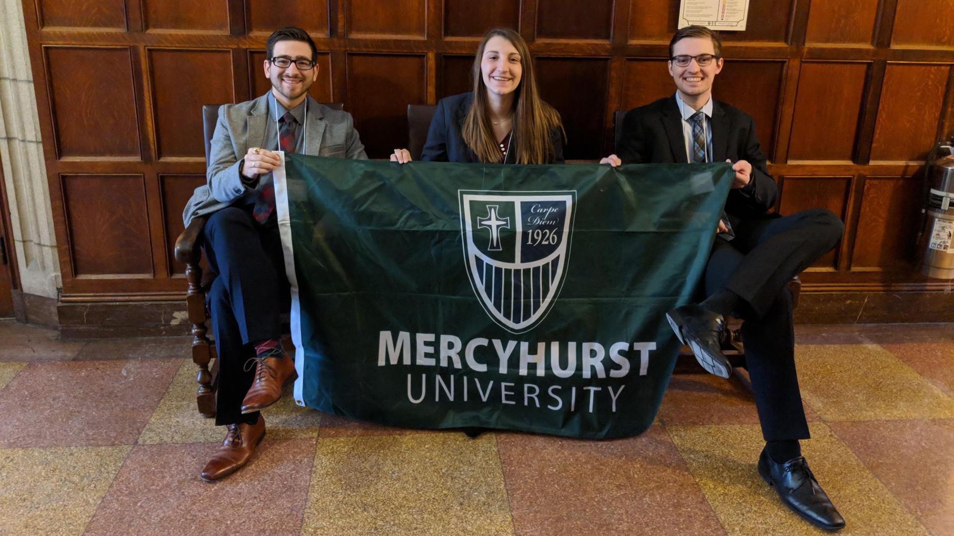 Religion and Society students sitting on a bench in formal attire holding the Mercyhurst Flag