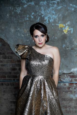 Broadway star, singer, and actress Jessica Vosk posing for a picture in a gold dress in front of a brick wall