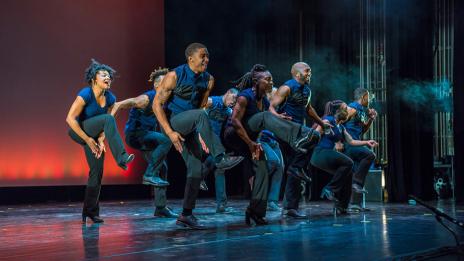 Dance members of Step Africa!, clapping under their lifted legs on a stage with blue and red lighting